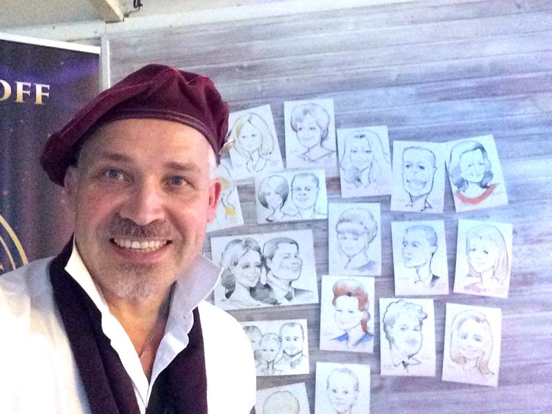 Шаржист Мишель позирует на фоне выставки своих шаржей в кафе 
